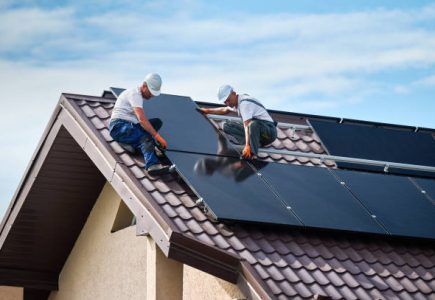 Workers building solar panel system on rooftop of house. Two men installers in helmets installing photovoltaic solar module outdoors. Alternative, green and renewable energy generation concept.
